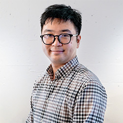 A portrait photo of a man wearing a checkered shirt and glasses, smiling and facing the camera against a plain background.