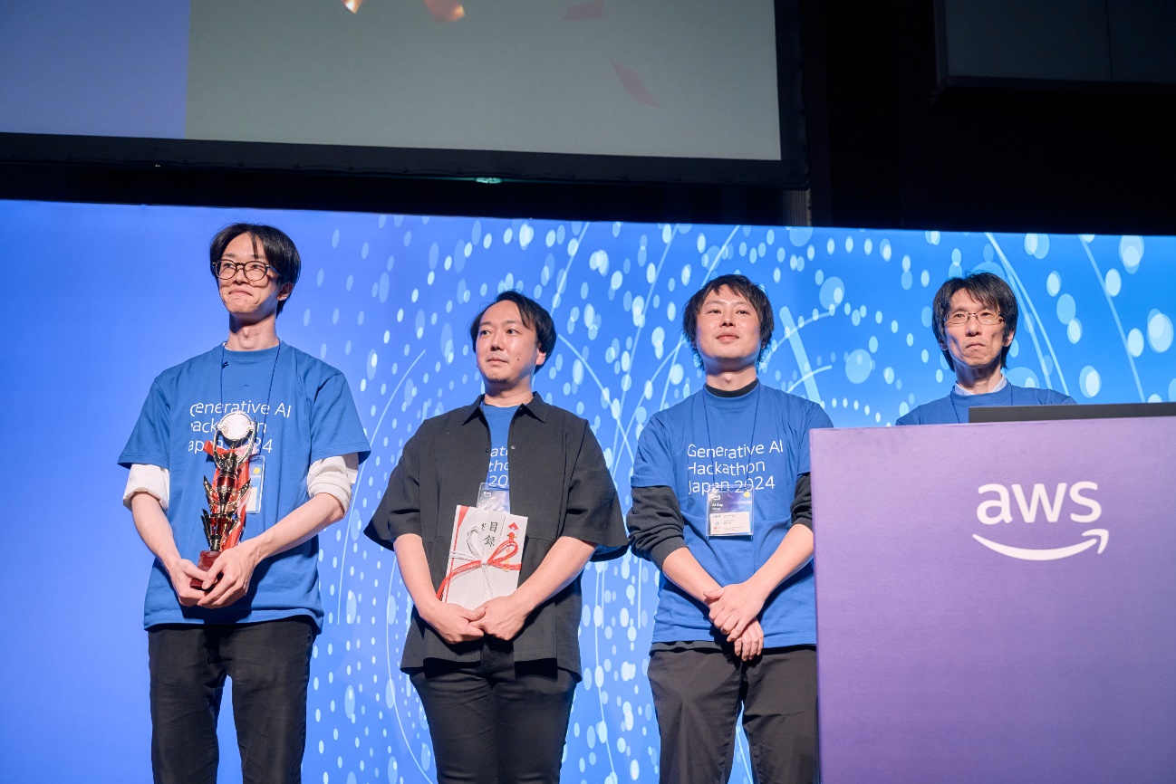 Group of participants standing on stage at the Generative AI Hackathon Japan 2024 award ceremony, wearing event T-shirts and holding awards, with an AWS-branded podium in front of a blue backdrop.
