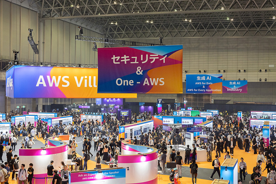 A large exhibition hall with colorful banners displaying "AWS Village" and "One - AWS," surrounded by booths and a crowd of attendees.