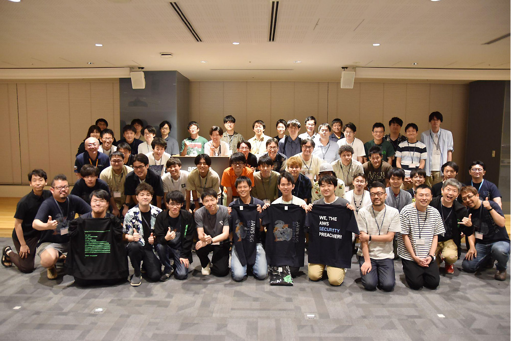 Large group photo of participants at AWS Security JAWS DAYS 2023 event, featuring attendees standing and sitting together, some holding shirts with event messaging, in an indoor conference setting.