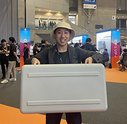 A person wearing a hat is holding a large case at an event booth. The background shows other attendees and exhibition stands in a convention center setting.