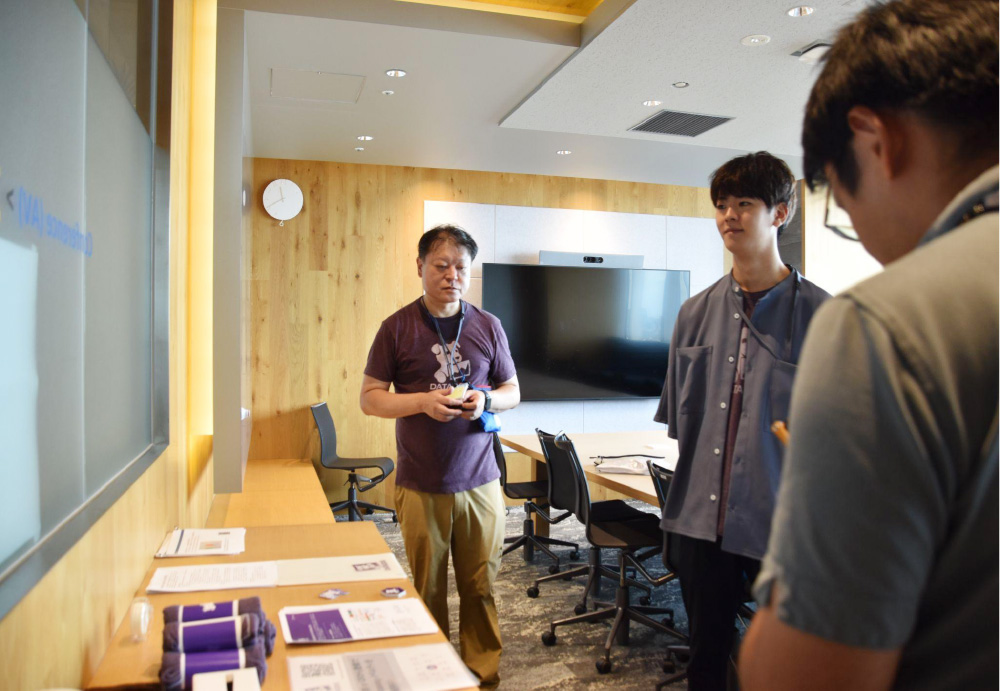 A group of people participate in a discussion in a modern meeting room during the JAWS DAYS 2023 security event. Conference materials and event items are laid out on a table, and a large screen is visible in the background.