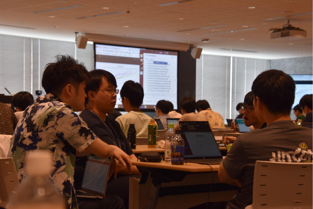 A group of participants engaged in a technology workshop, discussing around tables with laptops, bottles, and a projector screen displaying software in the background.