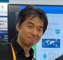 A person standing in front of a display booth at a tech event, with screens displaying informational graphics in the background.