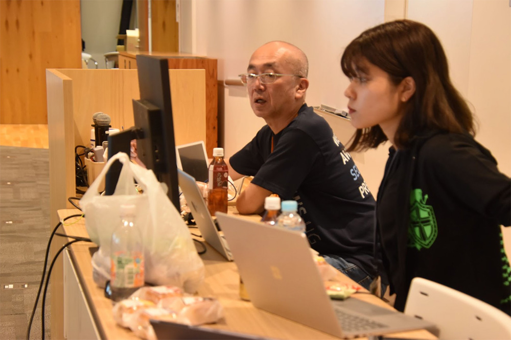 Two participants working at laptops and monitors during the JAWS Days security event, with drinks and snacks on the desk.