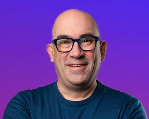 A bald man wearing dark-rimmed glasses and a blue shirt, smiling in front of a purple gradient background.