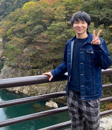 A person standing on a bridge making a peace sign, with a scenic forest and river in the background.
