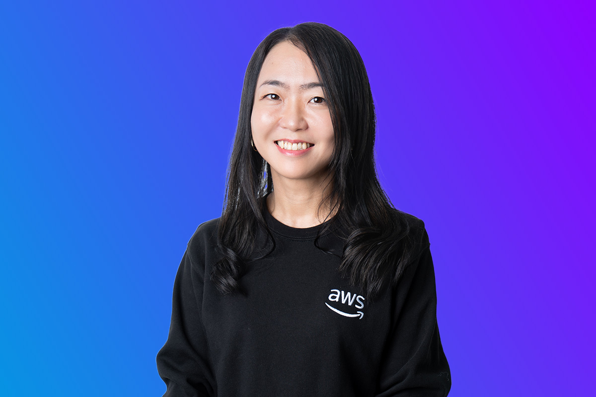 A woman wearing an AWS-branded black sweatshirt, smiling in front of a blue and purple gradient background. Studio portrait for AWS Japan Summit event materials.