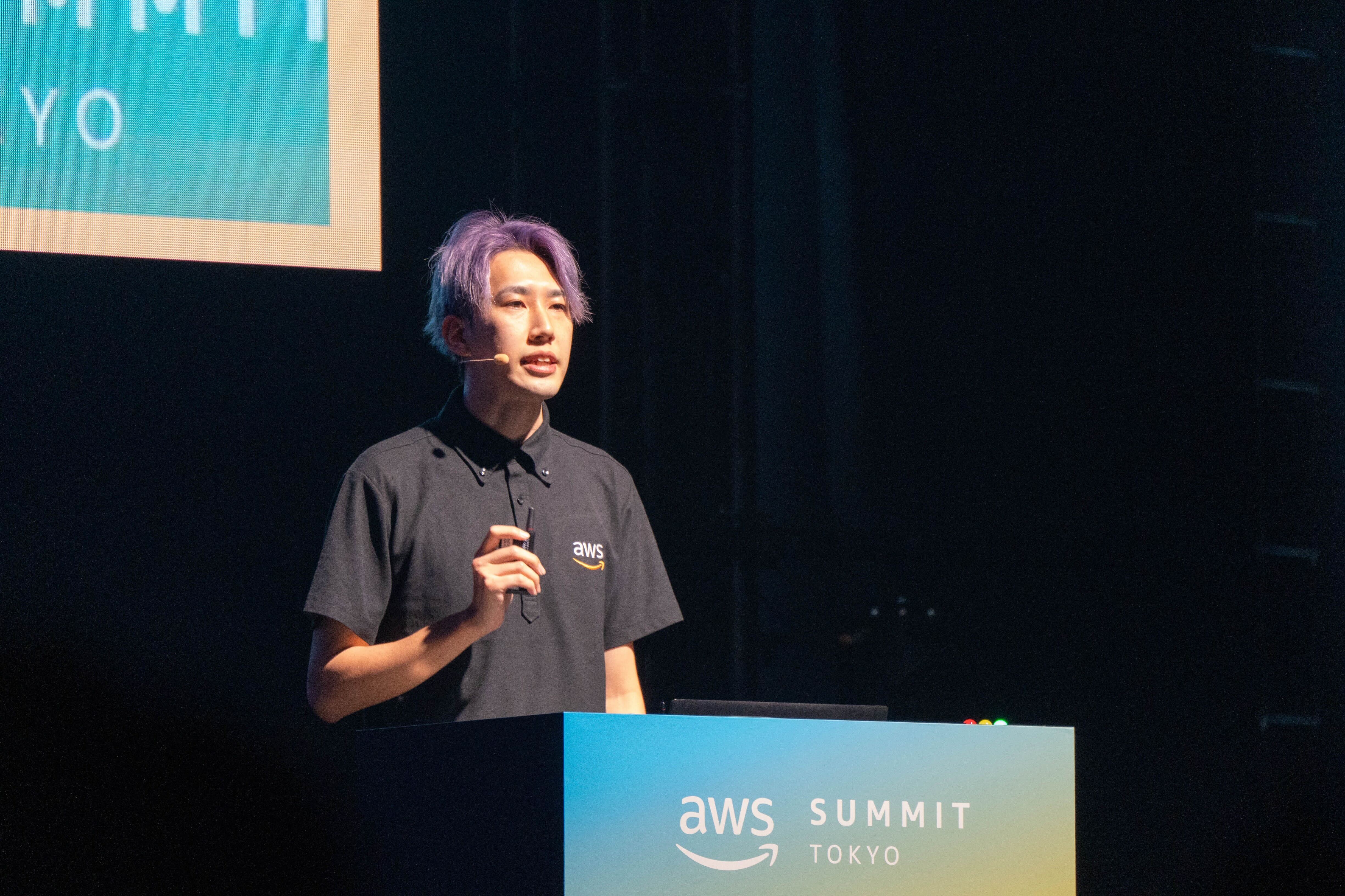 A person stands at a podium labeled 'AWS Summit Tokyo,' delivering a presentation on stage with a microphone headset. The background includes event signage and dark lighting, highlighting the speaker.