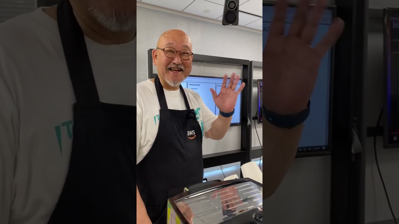 A friendly barista wearing an AWS-branded apron waves and smiles in a modern event space, standing next to a coffee machine. This image is used as a YouTube video thumbnail.