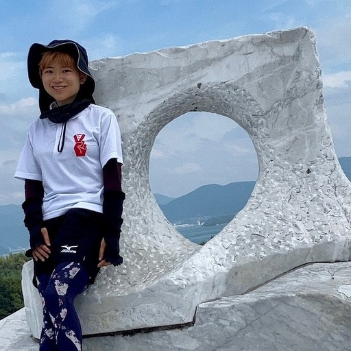 A person standing and smiling on a mountain summit next to a large white stone sculpture with a circular opening, with scenic mountains and sky in the background.