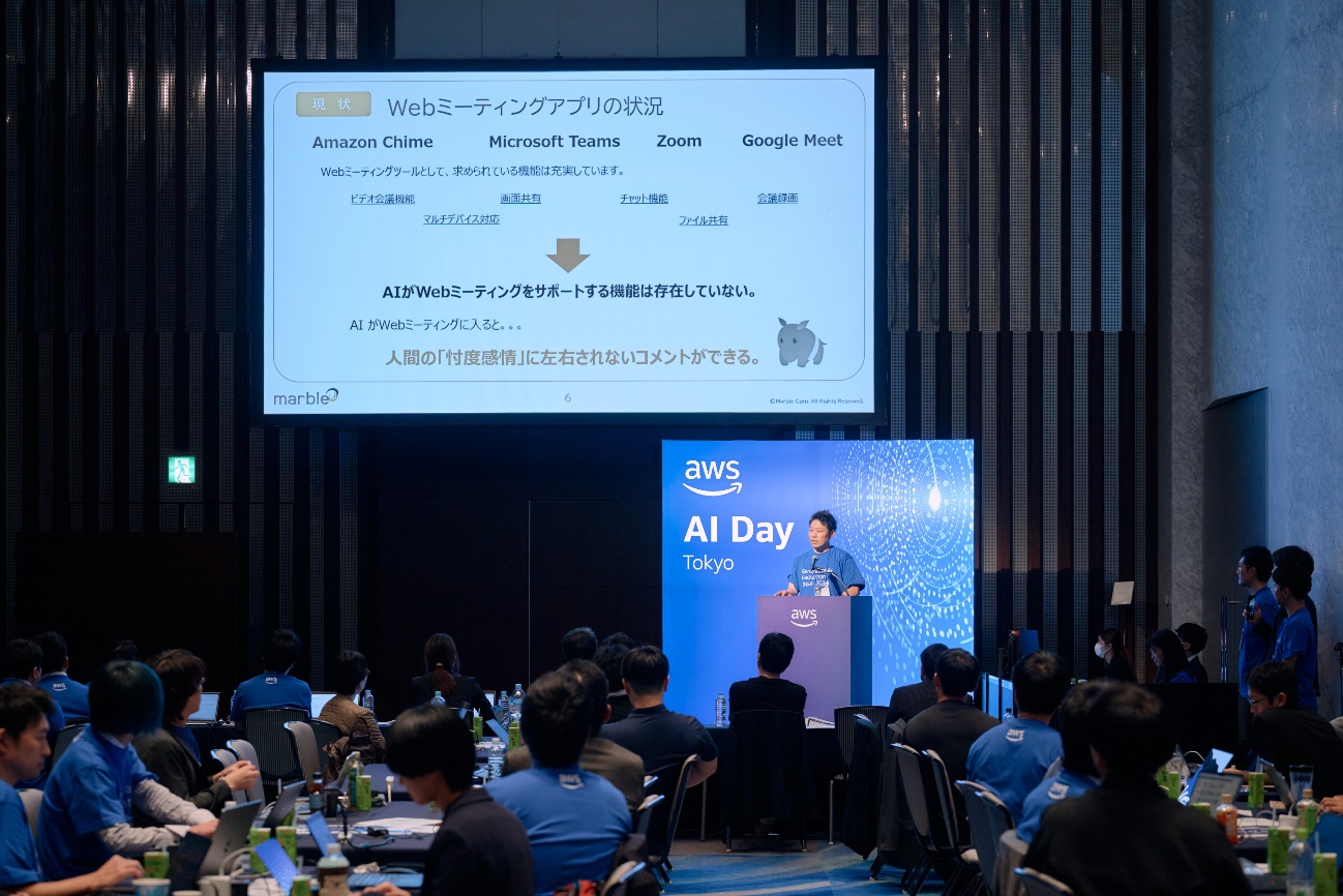 A presenter speaks at the podium during the AWS AI Day Tokyo 2025 hackathon, with participants seated and a large presentation slide displayed on screen. The event focuses on AI and cloud technology, highlighting collaboration, innovation, and web meeting applications.