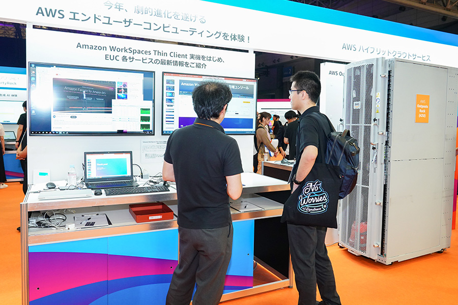 Two attendees observe a demonstration of Amazon WorkSpaces Thin Client and AWS Outposts at the AWS Summit Japan 2025. The exhibit features monitors displaying AWS End User Computing (EUC) services and a server rack labeled AWS Outposts, set within a technology event environment.