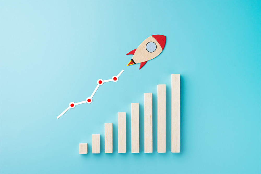 A wooden bar graph with increasing columns, a stylized paper rocket, and a red upward trend line symbolizing business growth and progress on a blue background.