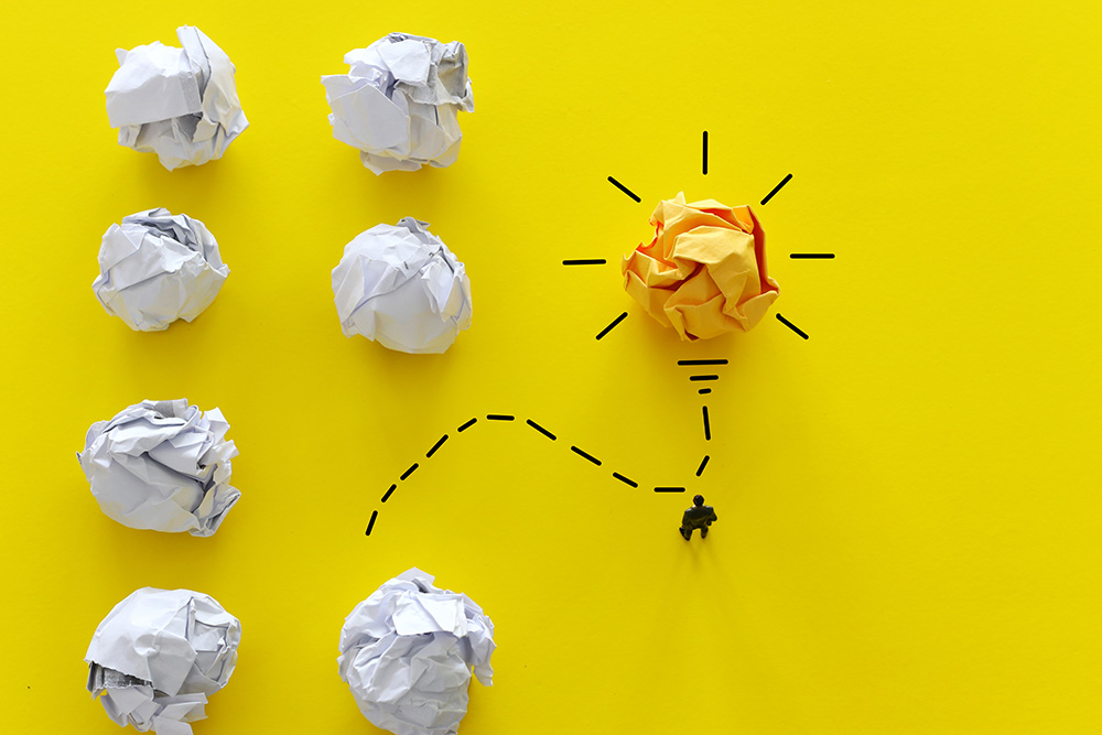 A conceptual image showing several crumpled paper balls on a yellow background, with one yellow ball depicted as a lightbulb and a small figure walking towards it, symbolizing creativity, problem solving, and new ideas.