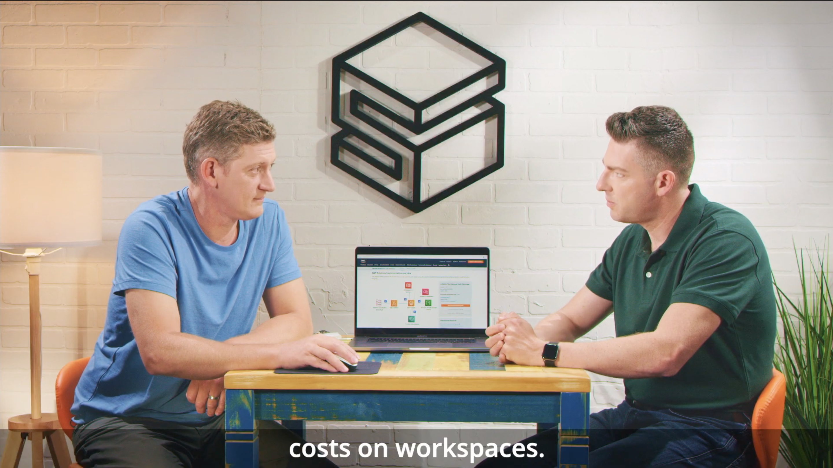 Two people sit at a table in a modern office setting, engaged in a discussion about AWS WorkSpaces. A laptop is open between them showing an AWS interface, and there is a large geometric wall logo behind them. Subtitles on the image read 'costs on workspaces.'