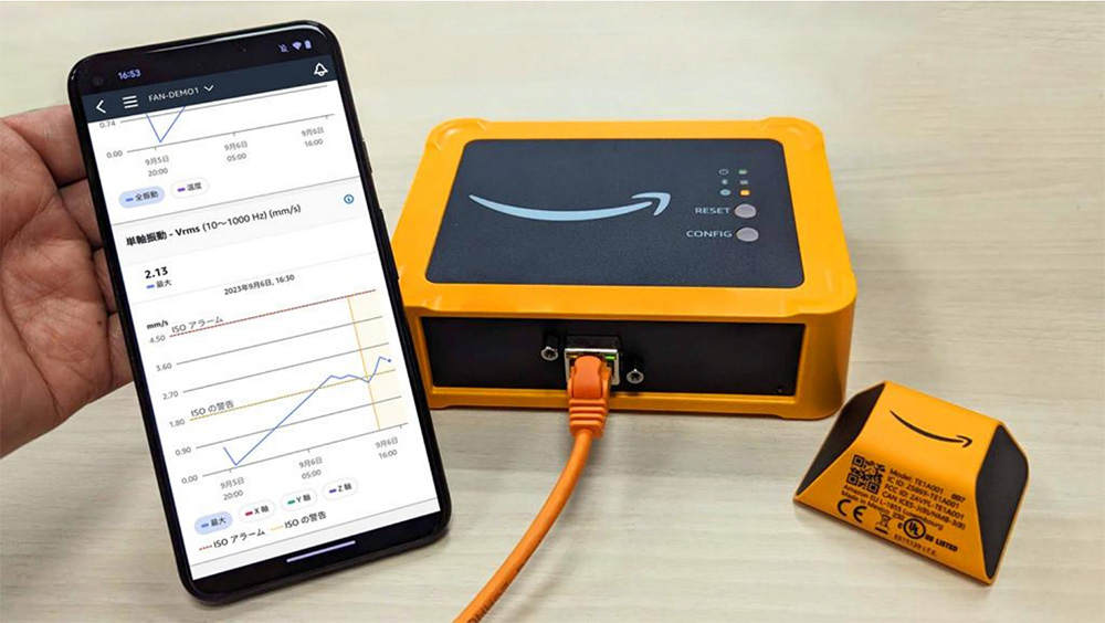 A photo of an Amazon Monitron device setup, including the gateway, sensor, and a smartphone displaying the Monitron app dashboard with monitoring data and graphs.