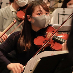 A woman wearing a face mask plays the violin in an orchestra. Sheet music and other musicians are visible in the background.