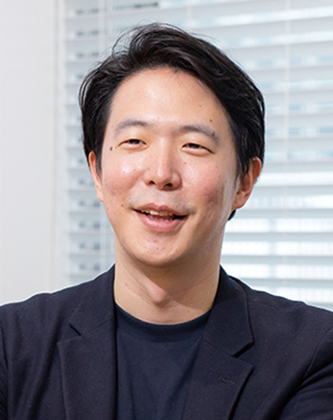 A portrait photograph of a man in a business casual outfit, identified as Mr. Shiohama from MICIN, against a background with window blinds.
