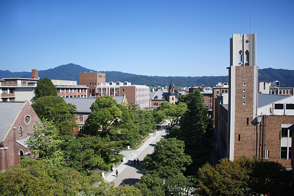 A scenic view of a university campus with brick buildings, a clock tower, tree-lined pathways, and mountains in the background under a clear blue sky.