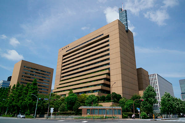 Exterior view of the Asahi Shimbun headquarters building in Tokyo, Japan, showing a modern multi-story office structure surrounded by greenery on a clear day.