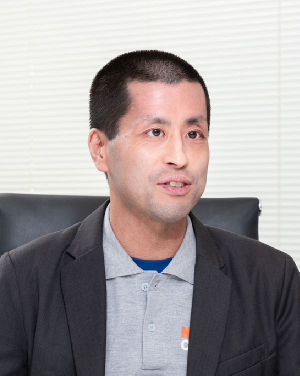 A person wearing a gray polo shirt with an orange logo and a dark blazer, seated in front of a white blind-covered window.