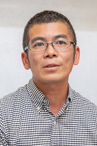 Portrait of Mr. Takahashi wearing glasses and a checkered shirt, facing forward against a neutral background.
