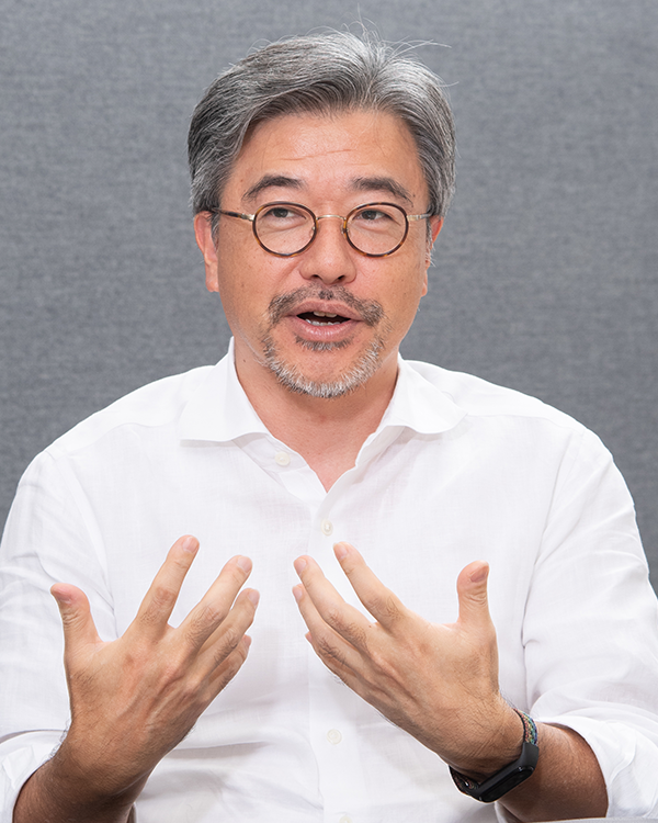 A portrait of an individual in a white shirt, wearing glasses, speaking and gesturing with hands, with a plain gray background. Used in the ExaWizards case study on the AWS Japan website.