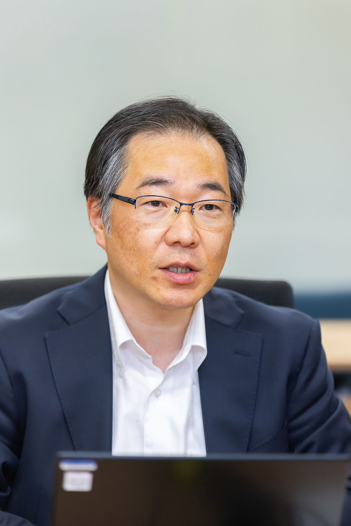 A portrait of a case study speaker from BIGLOBE, wearing glasses and a suit, seated at a desk in a professional setting.