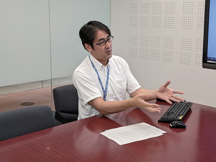A man in a white shirt and glasses is sitting at a conference table, gesturing as he explains something during an office meeting. There is a keyboard, mouse, and paper on the table in front of him.