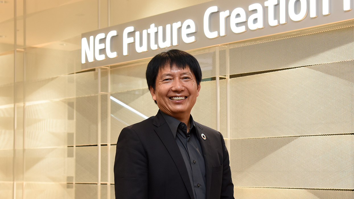 A smiling executive stands inside the NEC Future Creation Hall, with bright signage in the background. The setting conveys innovation and leadership in a modern business environment.