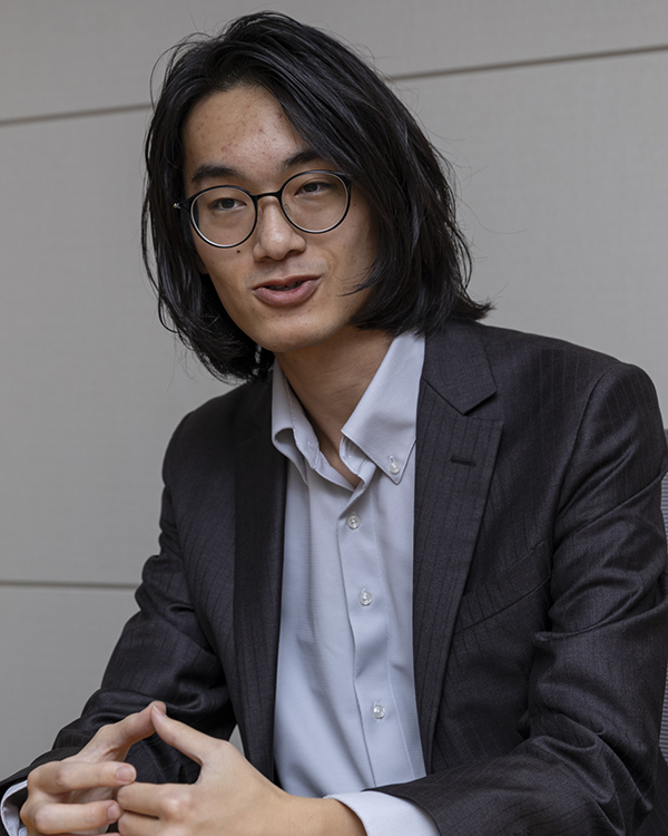 A person wearing a dark suit and light-colored shirt, sitting with hands clasped.