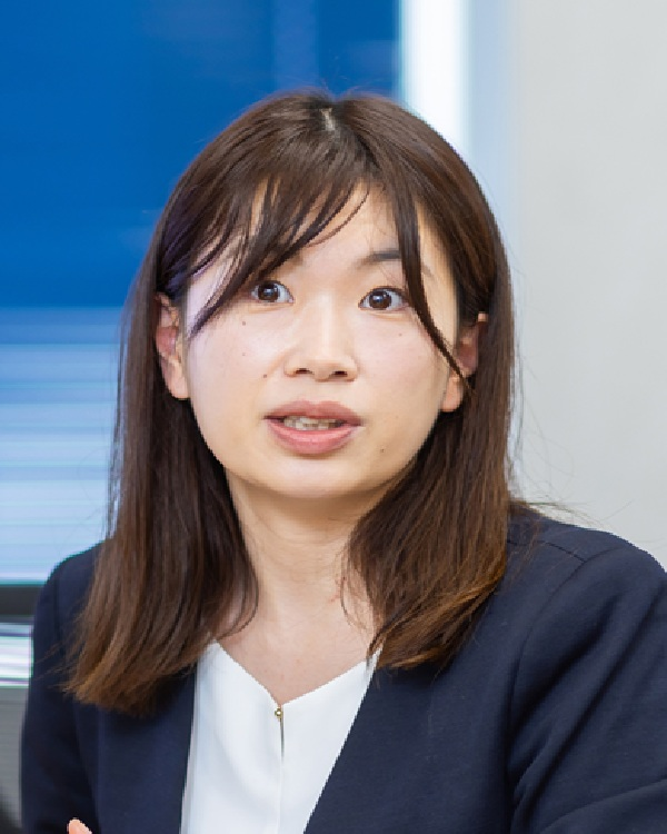 A professional portrait of a woman, used in the Yasashii-te Takagi case study. She is seated indoors, wearing a dark jacket and white blouse, appearing engaged in conversation.