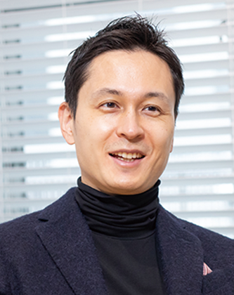 Portrait photo of a man wearing a dark blazer and turtleneck, sitting indoors in front of window blinds.