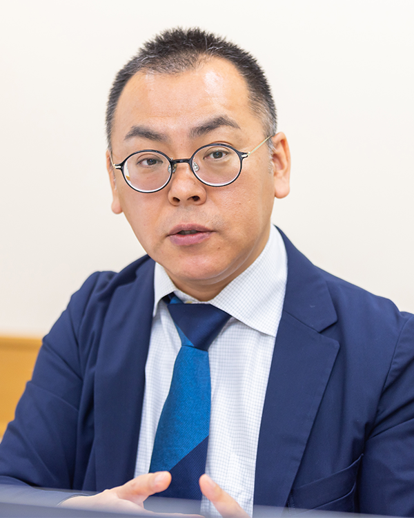 A business professional in a blue suit, glasses, and white shirt is speaking during an interview.