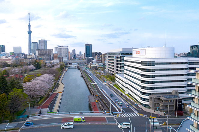 LIXIL headquarters building beside a river with cherry blossoms in full bloom, expansive Tokyo skyline, and Tokyo Skytree visible in the background.