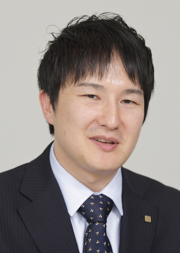 A person wearing a dark suit, light blue shirt, and patterned tie with a lapel pin.