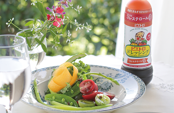 A plate of fresh vegetables and salad with a bottle of Pietro salad dressing on a table, accompanied by a glass of water and flowers in the background.