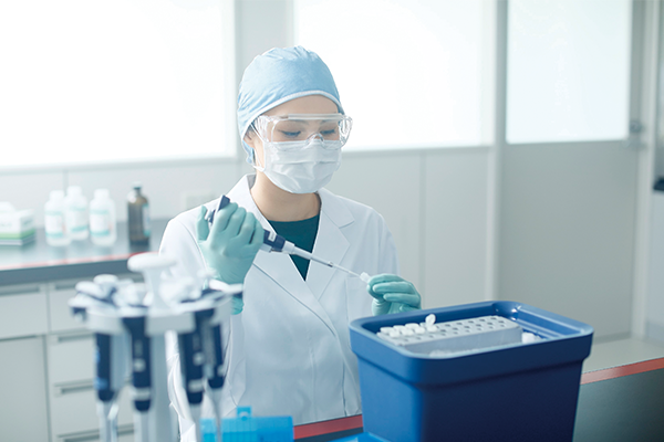 A scientist in a lab coat and gloves using a pipette near a container of test tubes in a laboratory setting.