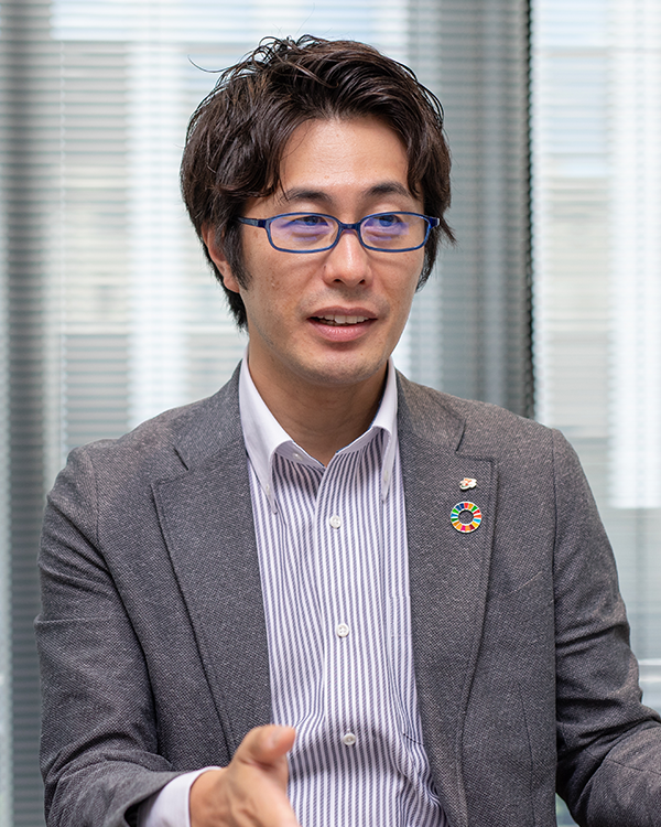 A businessman in a gray suit, striped shirt, and blue glasses, wearing an SDGs pin, giving an interview in an office setting.
