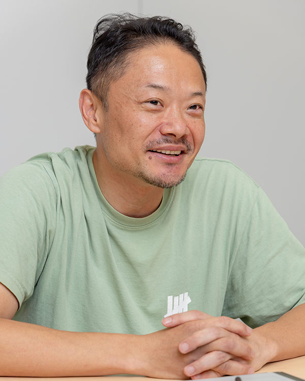 A person wearing a light green T-shirt with a white logo, sitting at a table with hands clasped.