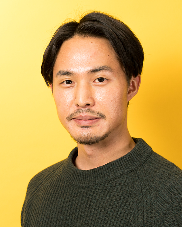 A portrait of a man with dark hair and a short beard, wearing a dark sweater, standing in front of a yellow background.
