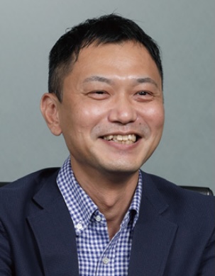 A person wearing a navy blazer over a checkered shirt, seated against a plain background.