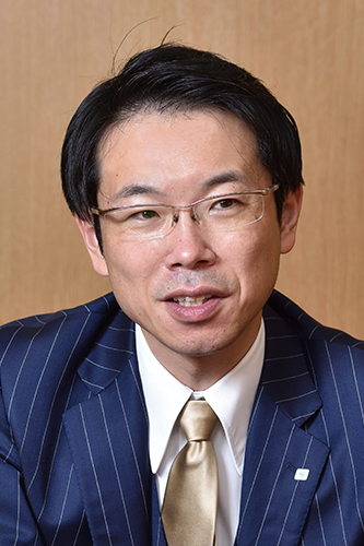 Portrait of an executive wearing a blue pinstripe suit, gold tie, and glasses, with a neutral brown background.