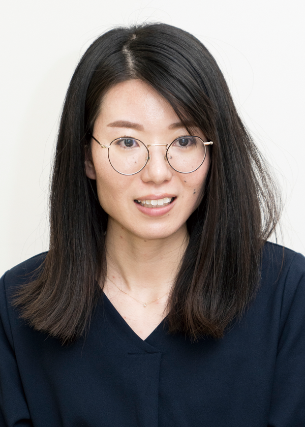 A portrait of a woman with long, straight dark hair wearing round glasses and a dark top, posing against a light background.