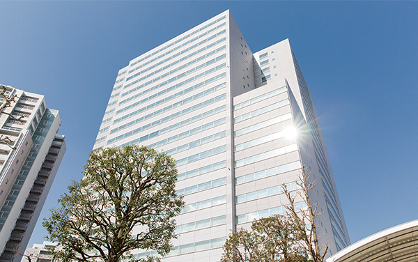 A modern high-rise office building in Tokyo, photographed from ground level with blue sky and trees in the foreground.