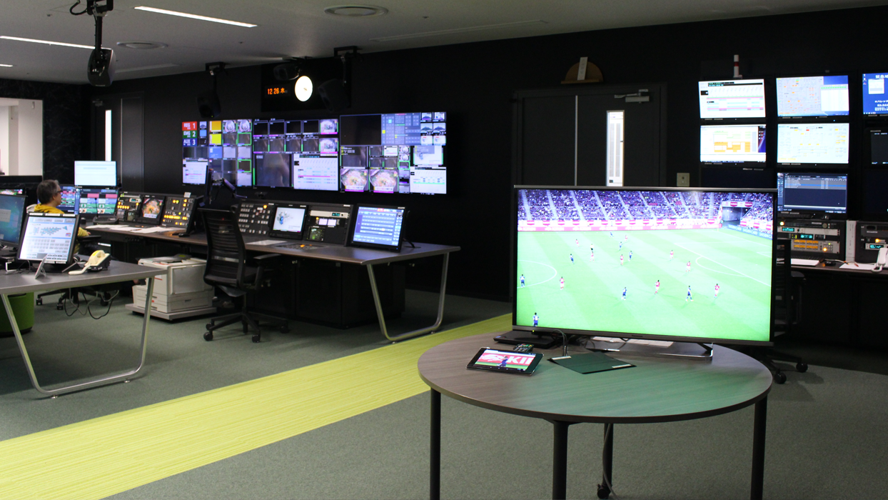 A broadcast control room with multiple monitors displaying various feeds, including a large screen showing a soccer match.