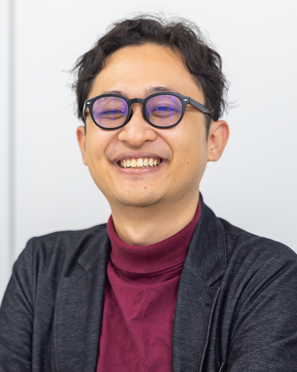 A smiling man wearing glasses, a maroon turtleneck, and a dark blazer.