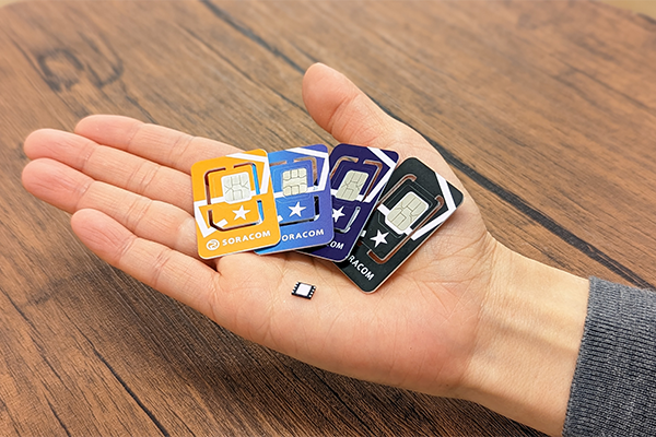 A hand holding several colorful Soracom SIM card holders and a small SIM card on a wooden surface.