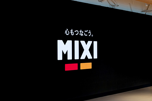 "Logo of Mixi with Japanese text above and red and orange bars below, displayed on a black wall."